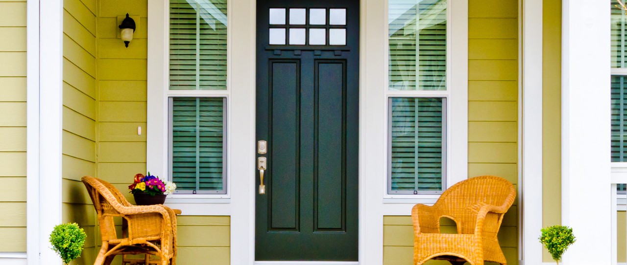 Yellow wooden home with gorgeous curb appeal and two woven chairs and a black door.